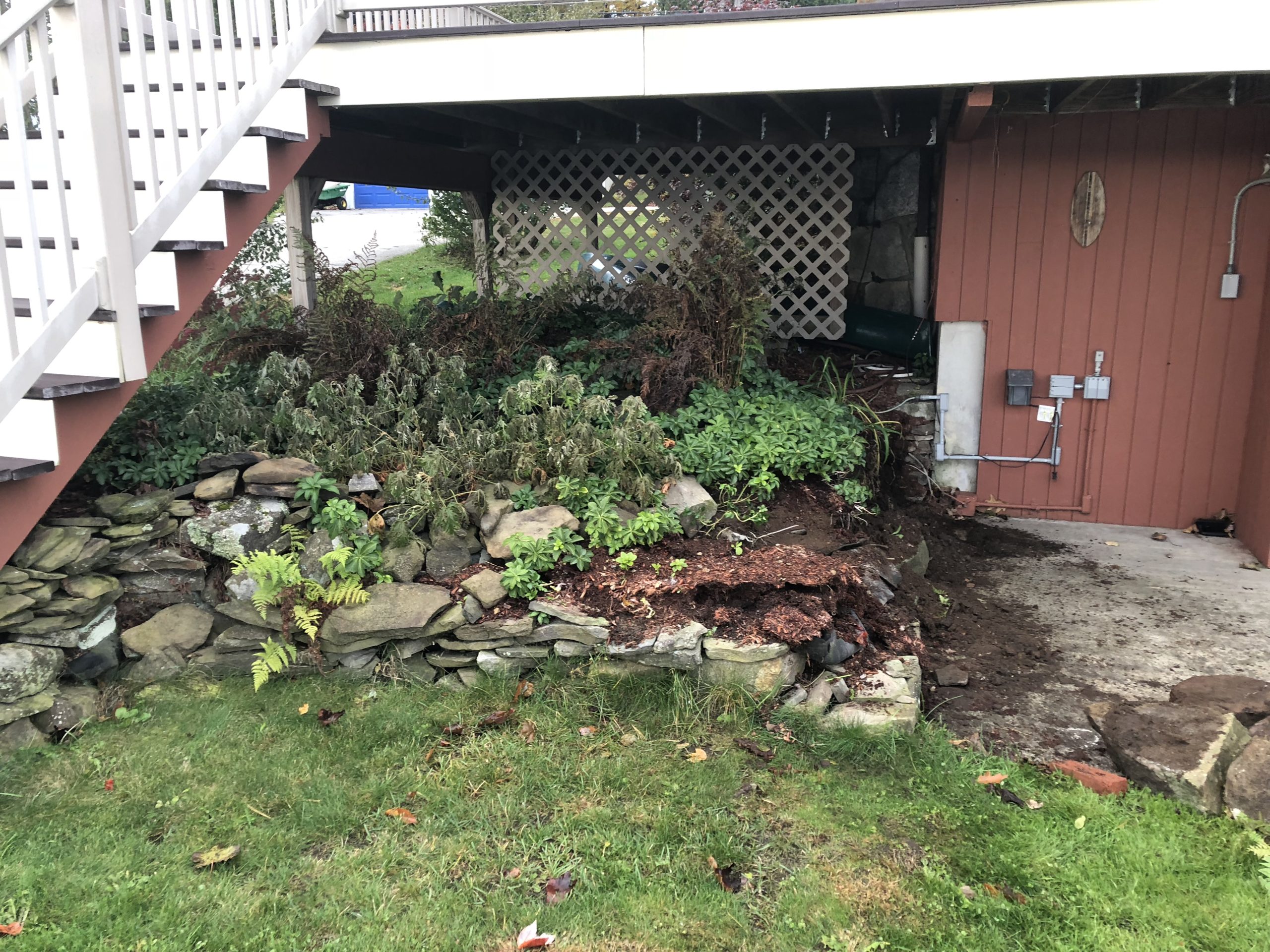 A wooden deck with stairs leads down to a garden area bordered by a stone wall. The garden is dense with green and brown plants, and the side of the building is visible with utility meters.