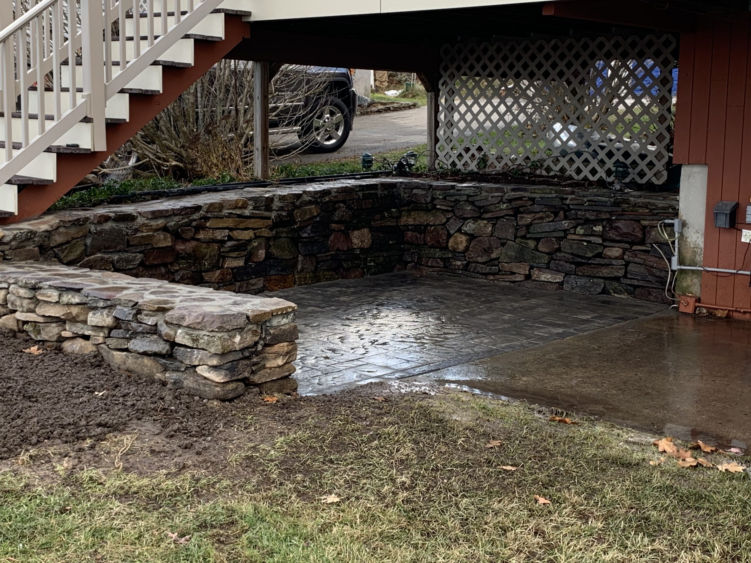 A stone retaining wall surrounds a paved patio area beneath a staircase and deck, with a vehicle visible in the background.