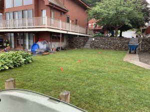 A neatly mowed lawn with red flags is in front of a two-story house. A stone wall borders one side, and a blue wheelbarrow is visible near the patio.