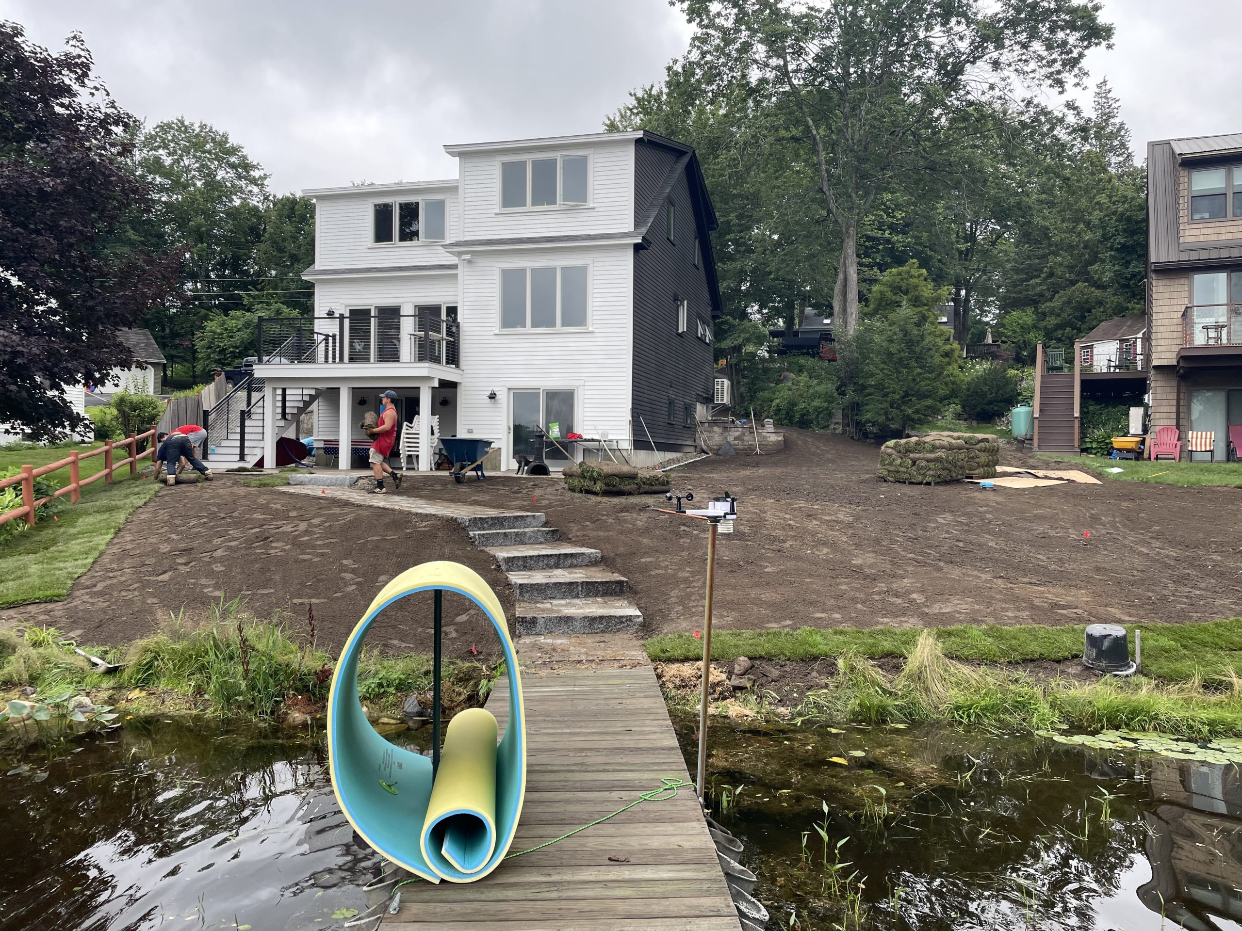 Backyard with newly laid dirt leading to a small pond, two people working near the house, and a rolled-up yoga mat on a dock. House is two stories with a deck.